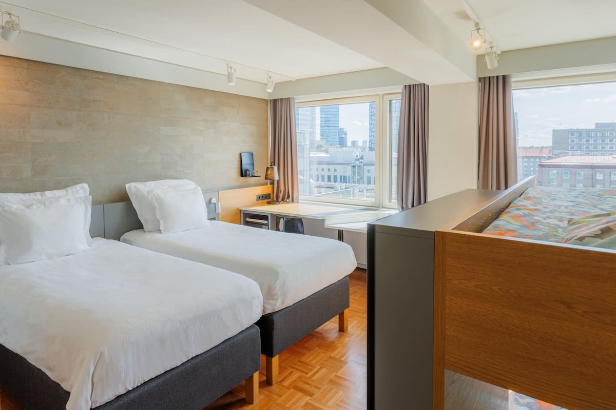 Une chambre d’hôtel avec deux lits, un bureau, une chaise, une lampe et une grande fenêtre donnant sur la ville.
