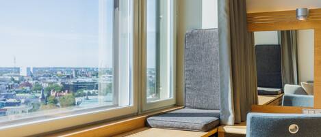 A hotel room with a large window offering a city view, a wooden desk, a cushioned bench, and a chair.