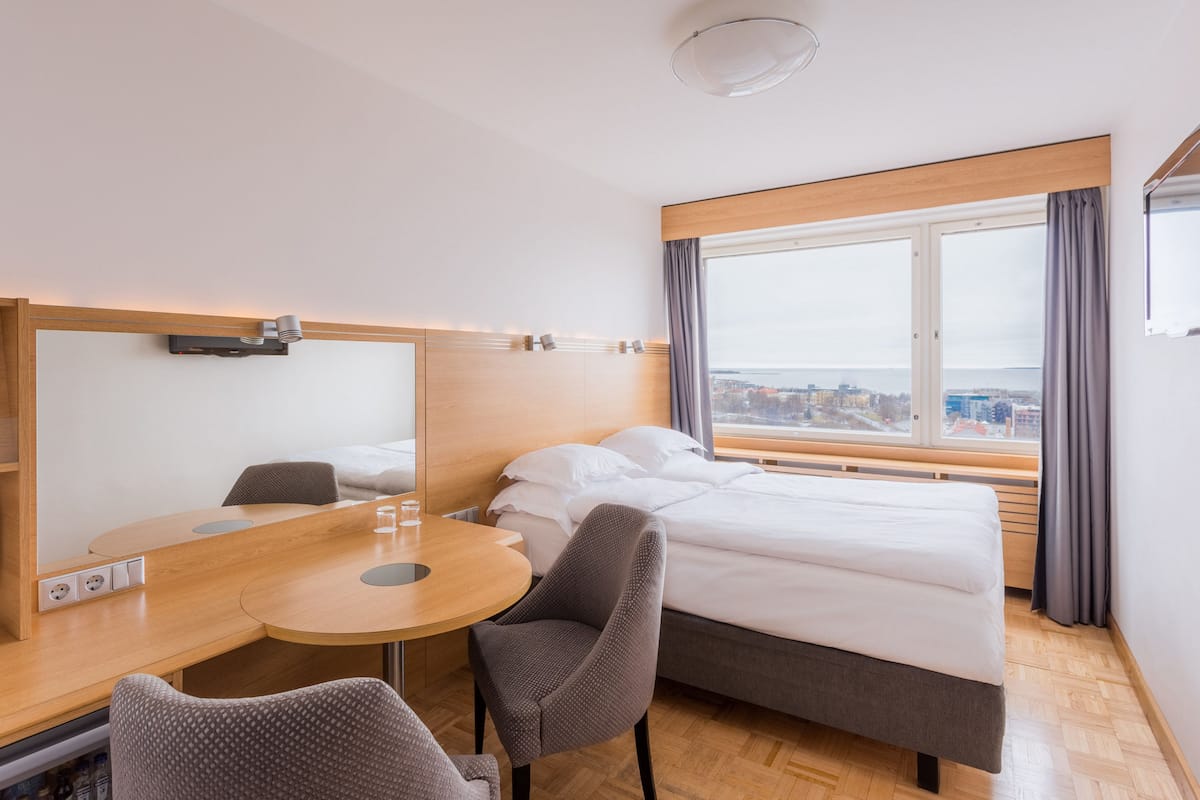 Une chambre d’hôtel avec un lit, un bureau et une chaise. Il y a une fenêtre donnant sur des immeubles et offrant une vue sur la ville.