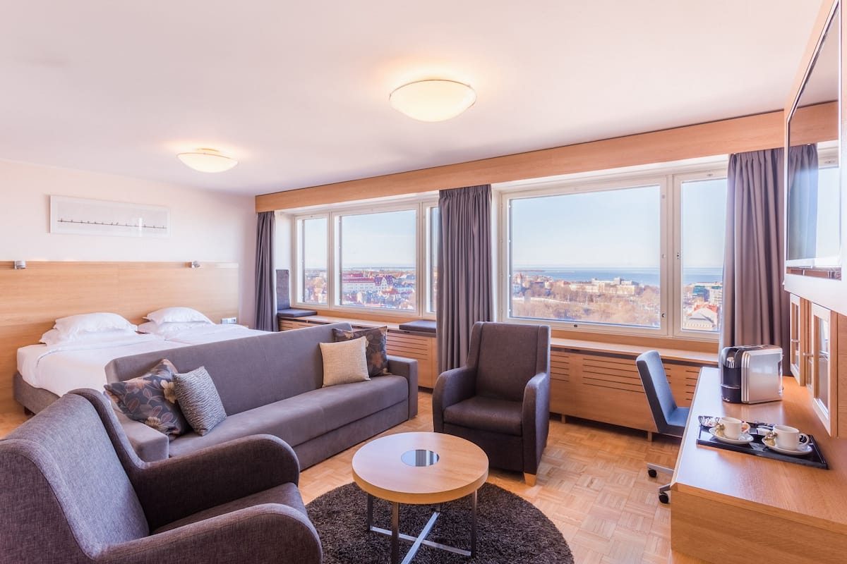 Une chambre d’hôtel comprenant un lit, un canapé, un fauteuil, une table basse et offrant une vue sur la ville grâce à de grandes fenêtres.