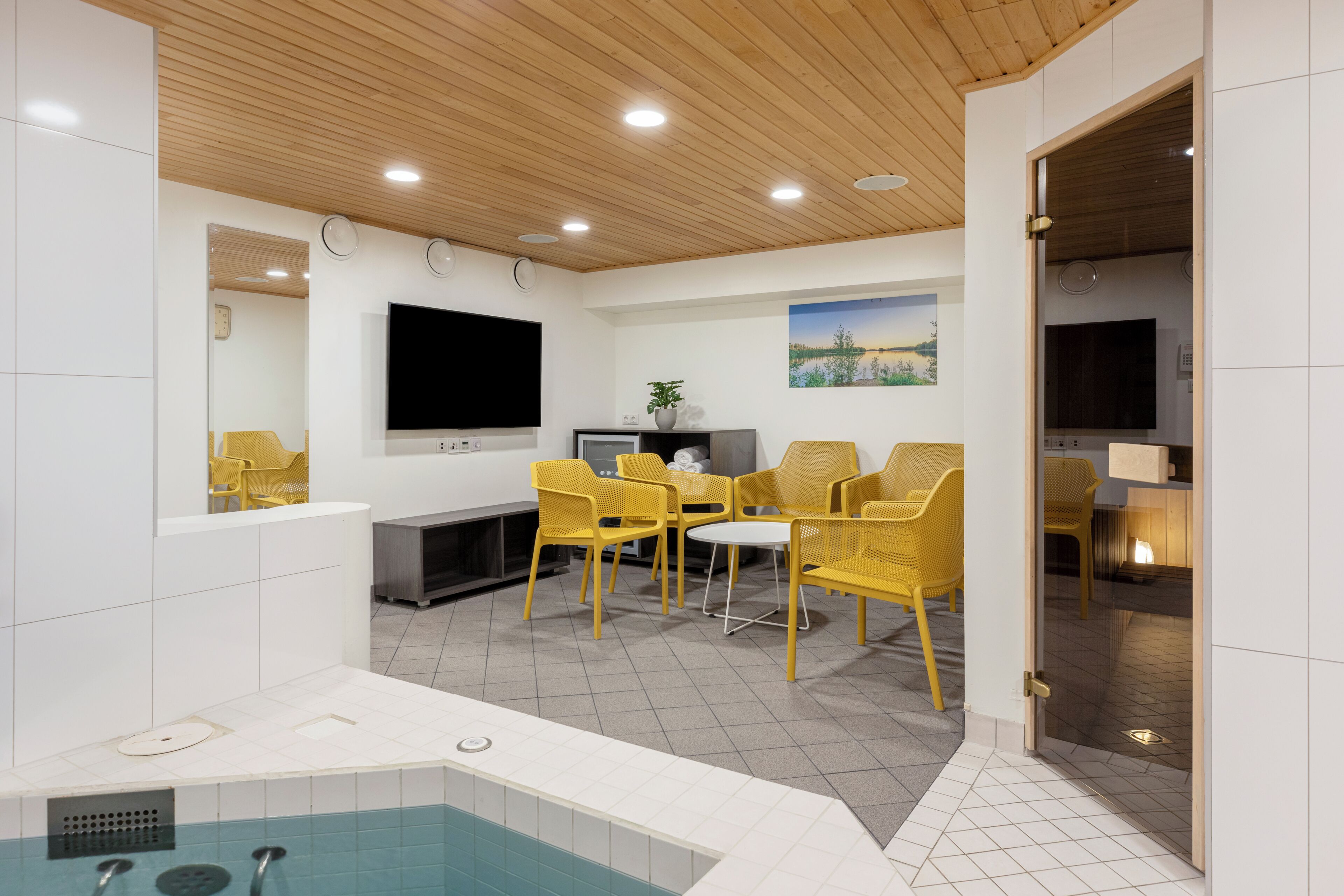 A modern indoor pool area with a wooden ceiling, a flat-screen TV, and yellow chairs.