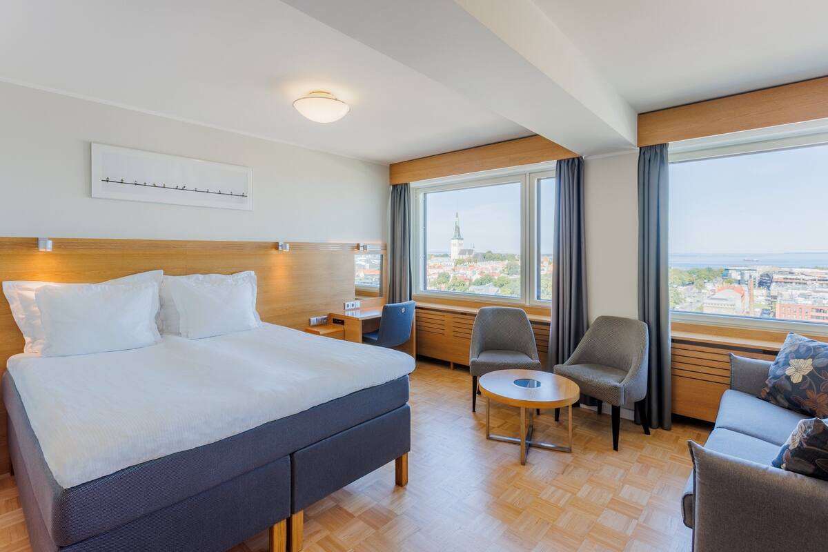 Une chambre d’hôtel avec un lit, deux chaises, une petite table et un canapé. Il y a une fenêtre donnant sur un paysage urbain.
