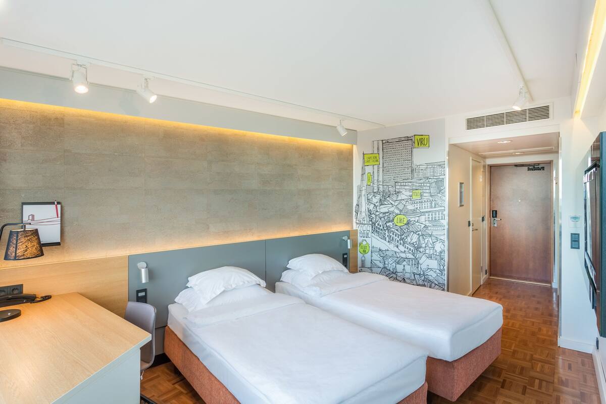 Une chambre d’hôtel moderne avec deux lits simples, un bureau et un mur décoré sur le thème des cartes géographiques.