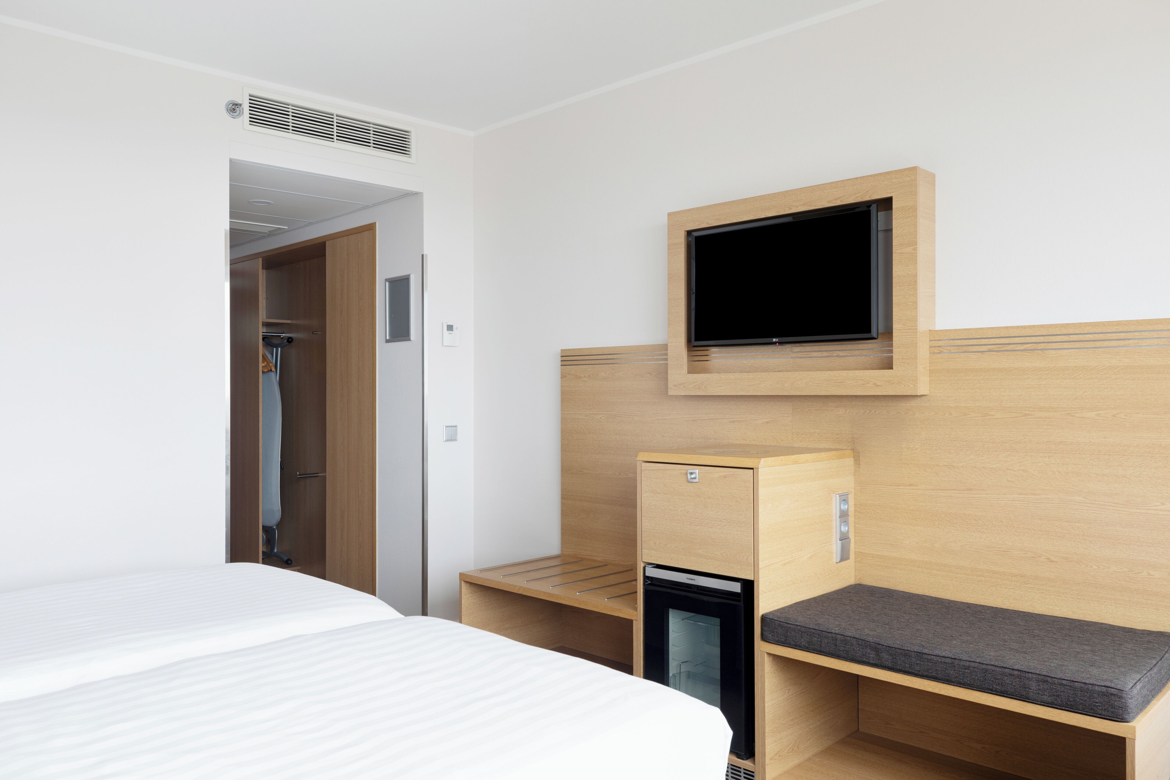 Quarto de hotel com cama, escrivaninha, minibar e televisão na parede.
