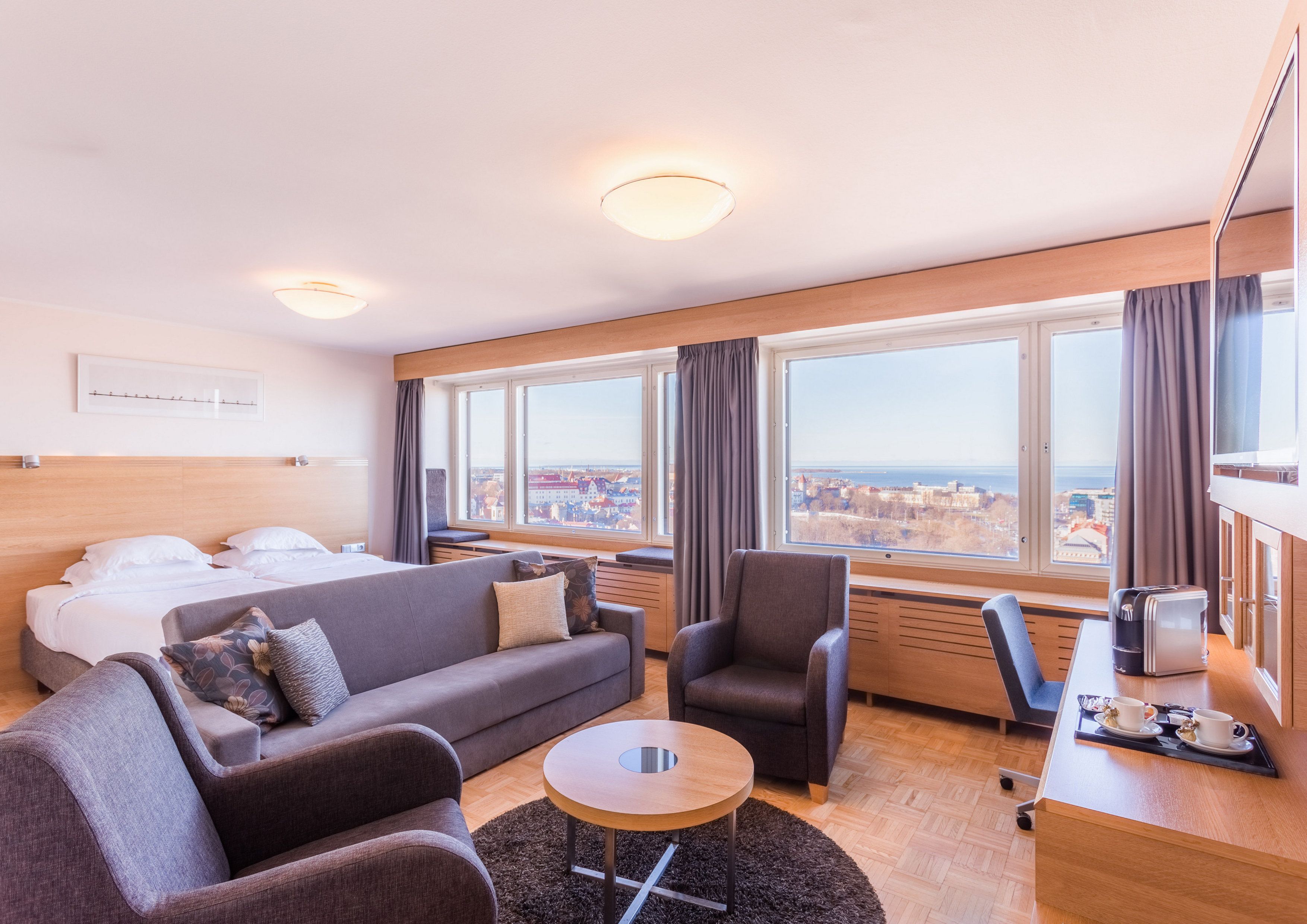 Habitación de hotel moderna con cama, sofá, sillón y una mesita. Amplios ventanales con vistas a la ciudad.