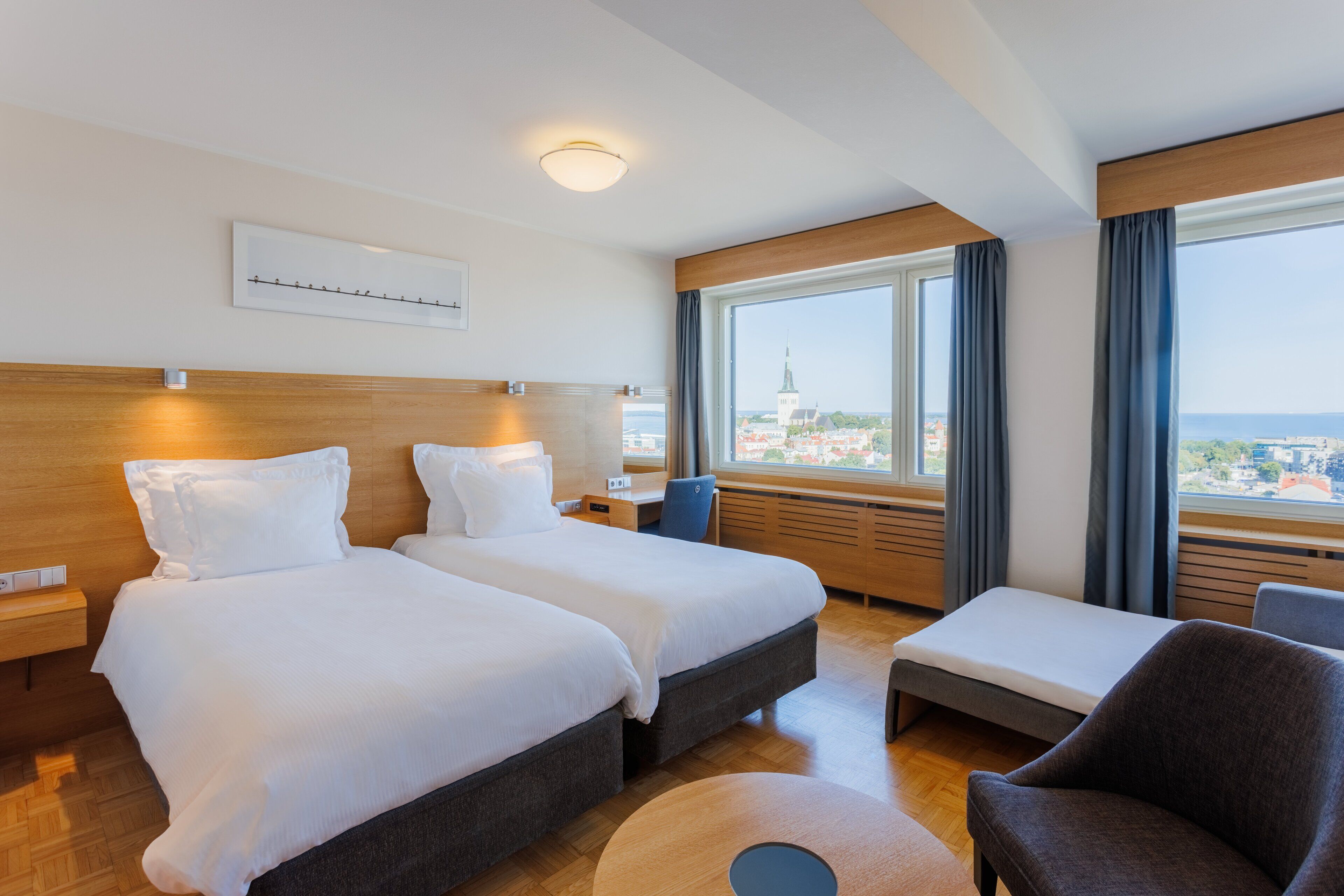 A hotel room with two beds, a desk, a chair, and a view of a cityscape through large windows.
