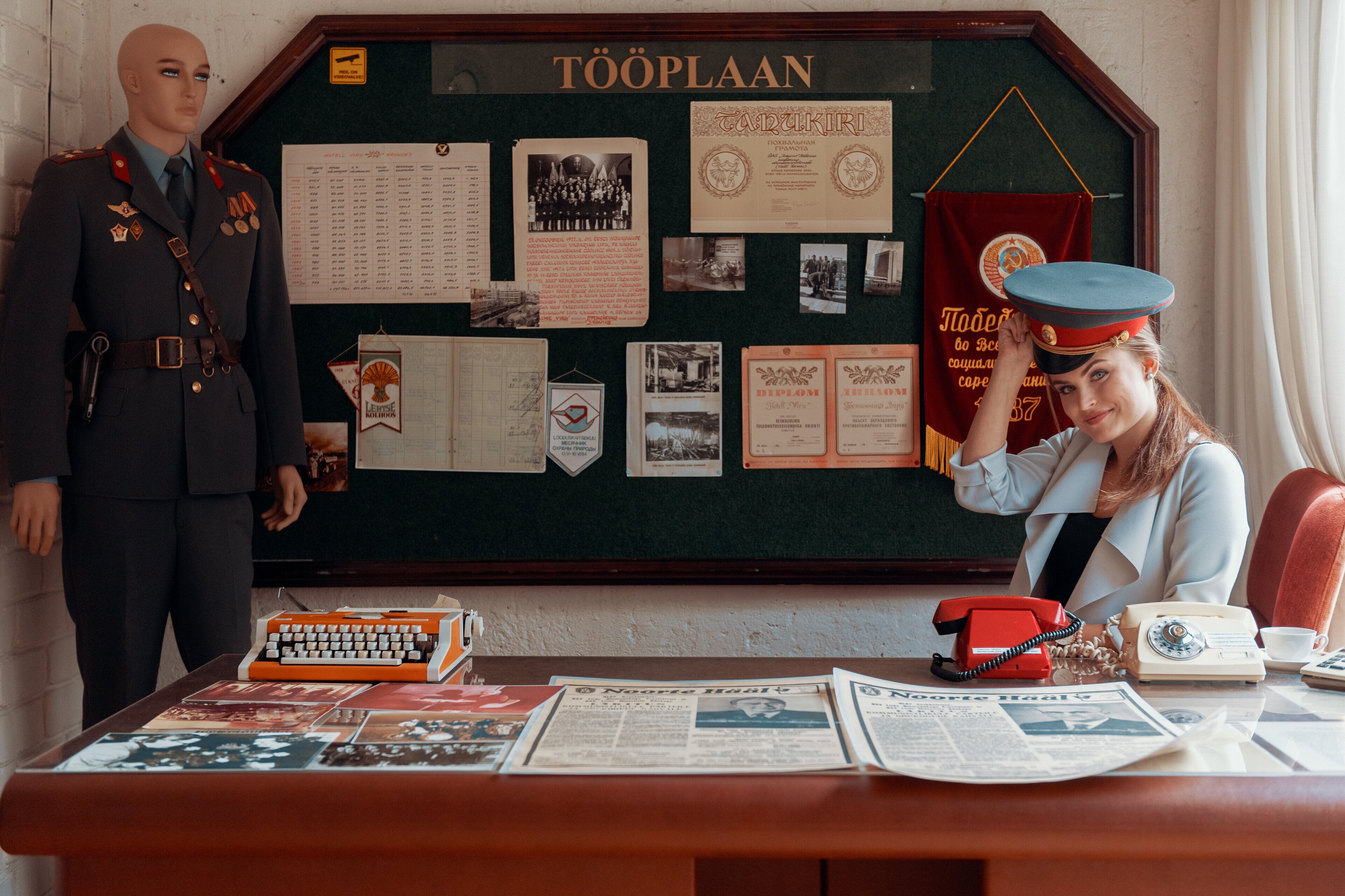 A room with a bulletin board displaying various papers and photographs, a mannequin in a uniform, and a desk with old-fashioned telephones and newspapers.