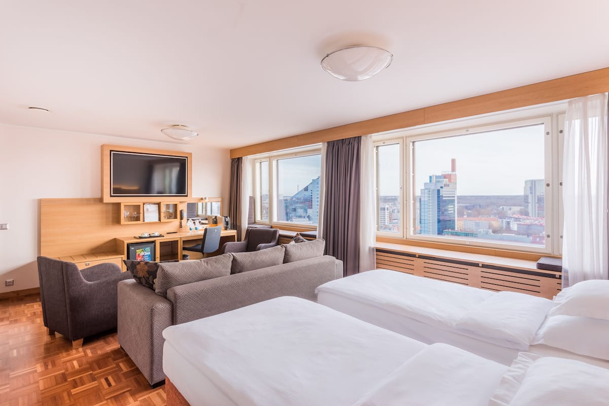 Une chambre d’hôtel comprenant un lit, un canapé, un bureau avec un ordinateur, une télévision et une grande fenêtre donnant sur la ville.