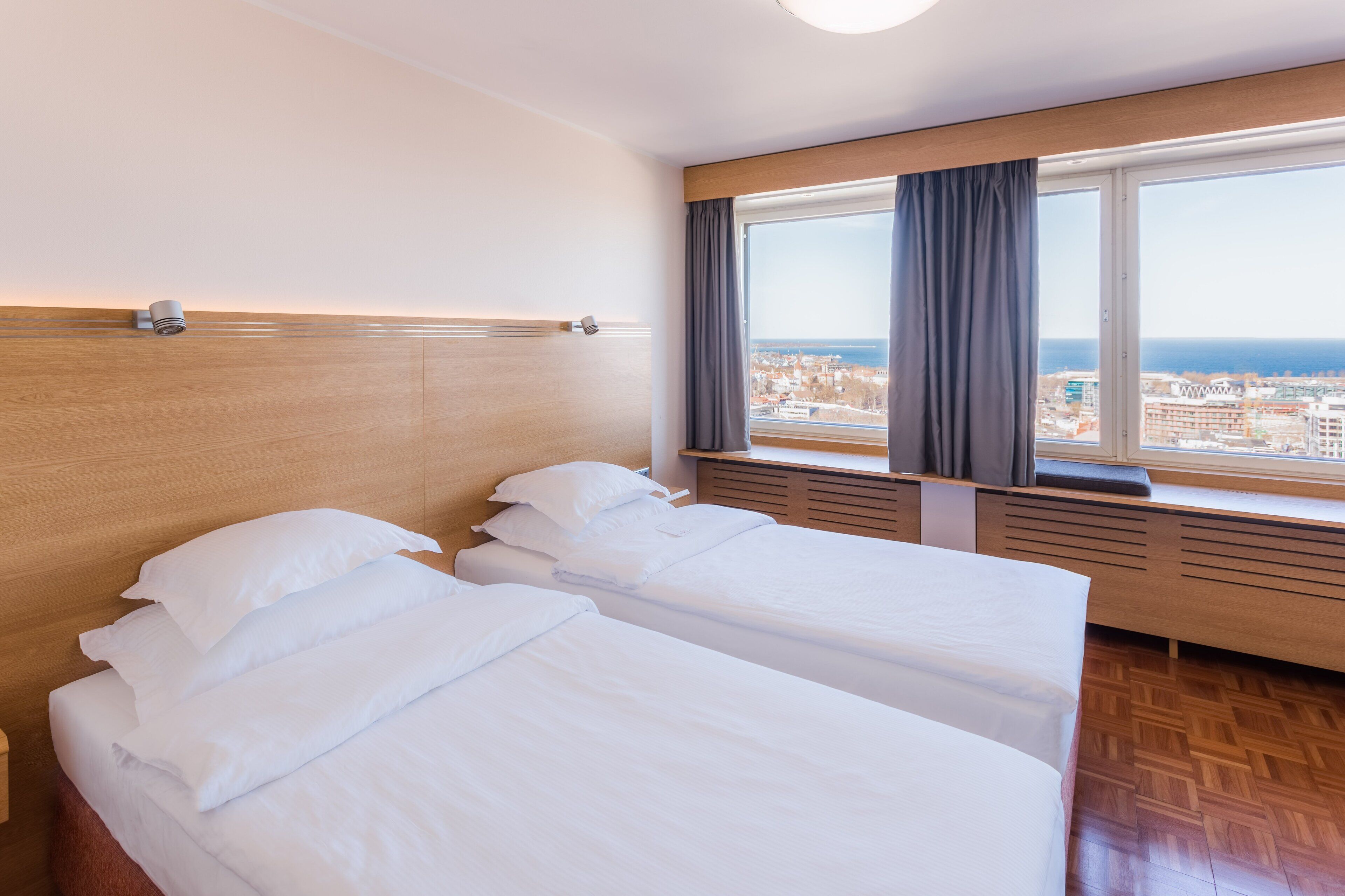 A hotel room with two beds, a large window with curtains, a view of the city, and a wooden headboard.