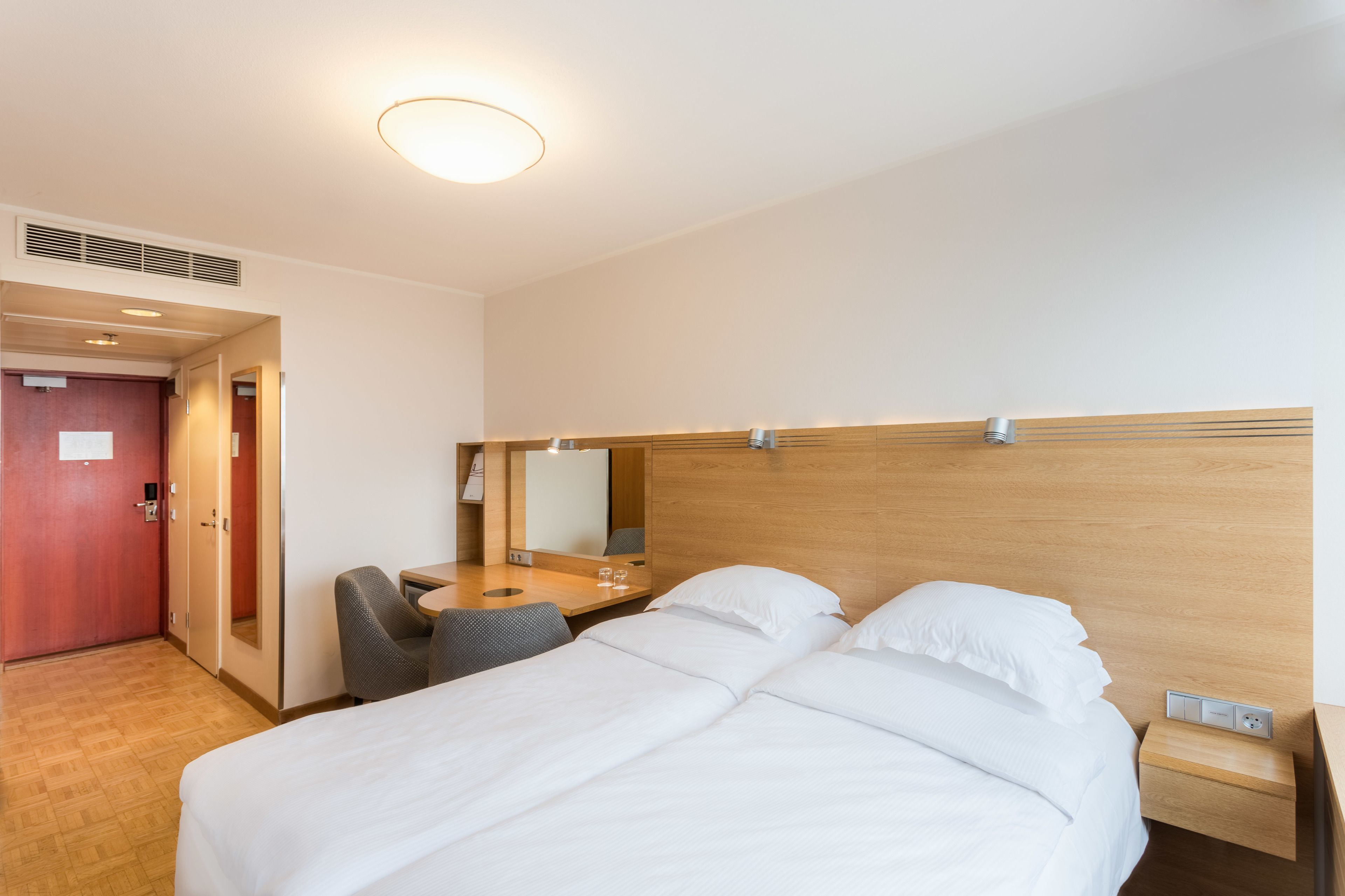 Una habitación de hotel con cama, escritorio, silla, espejo y cabecera de madera.