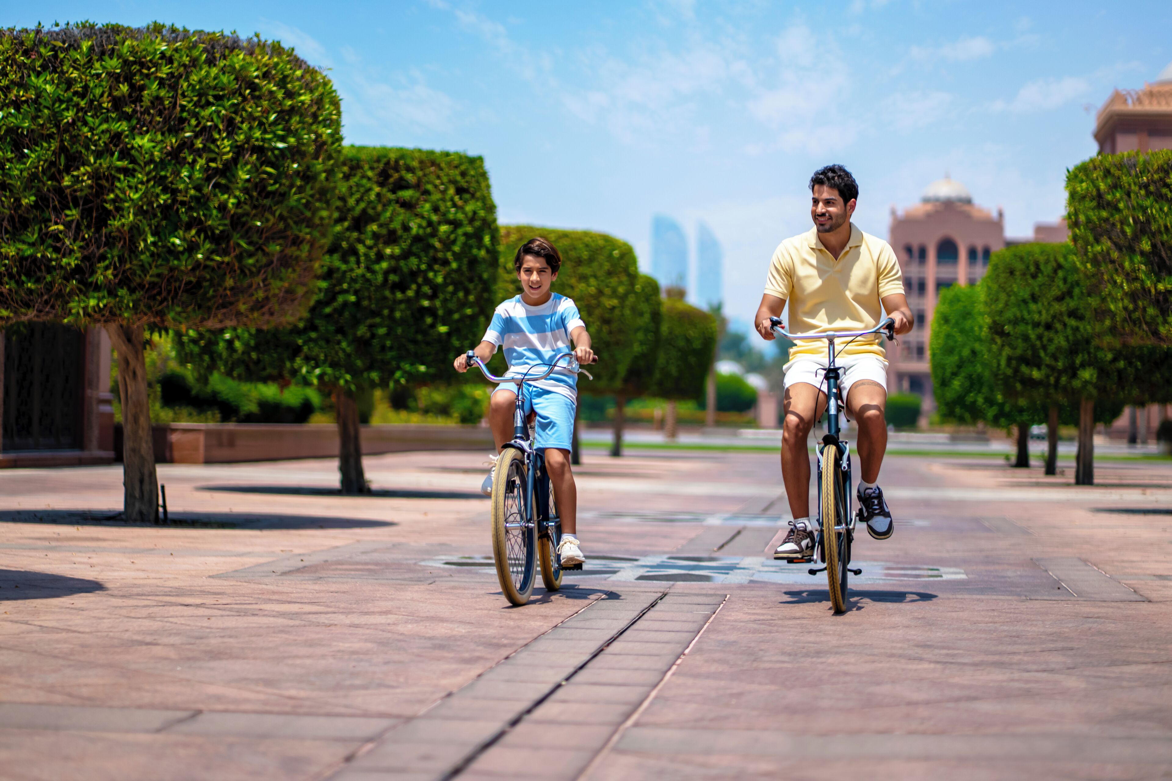Two people riding bicycles on a paved pathway with trimmed hedges and trees, in an urban setting with buildings in the background.
