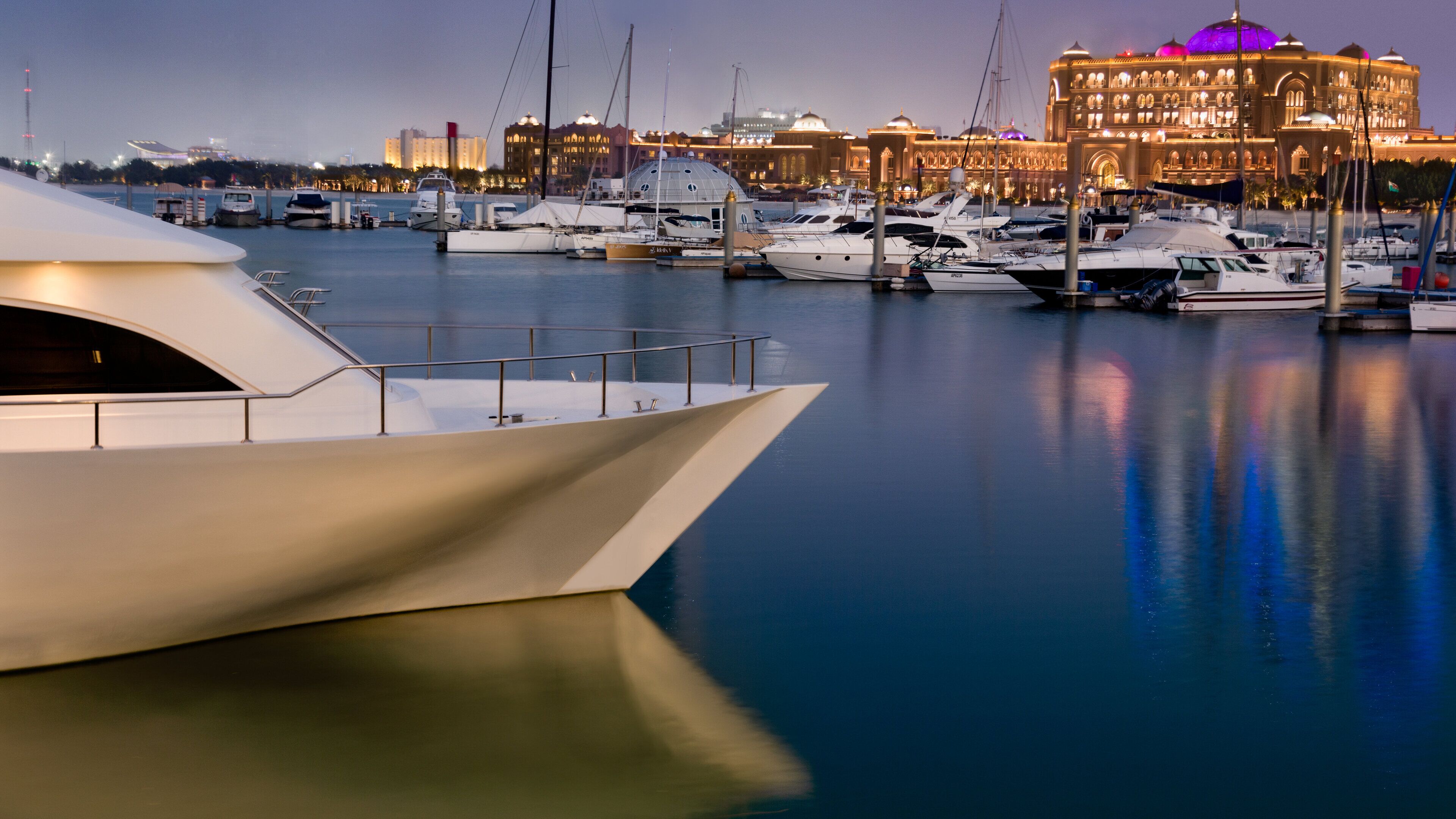 A marina with multiple yachts docked, a large illuminated building in the background, and calm water reflecting the lights.