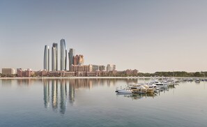 A marina with boats docked, a city skyline with modern buildings, and a calm body of water reflecting the structures.