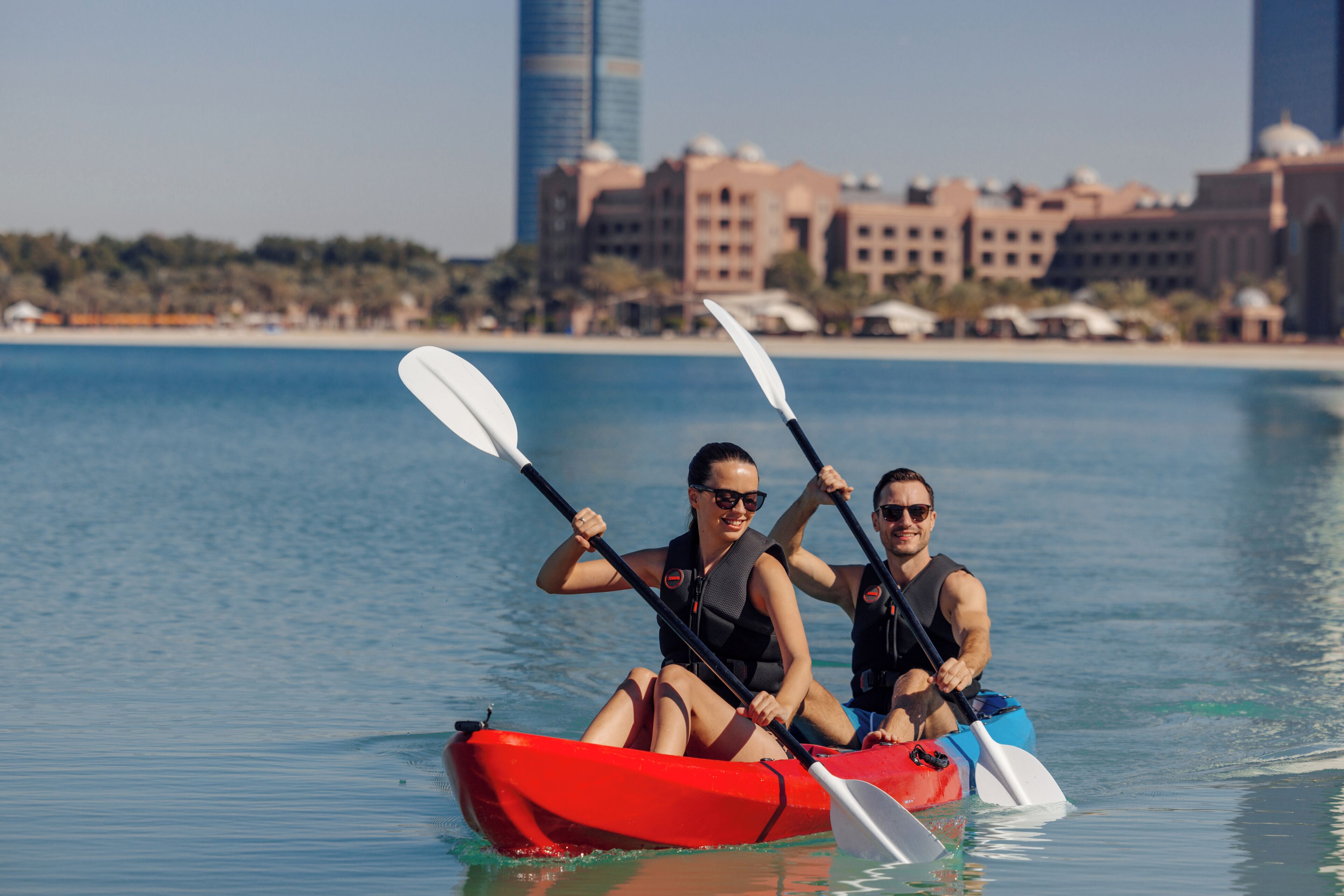 Two people kayaking in a red kayak on calm water with a cityscape in the background.