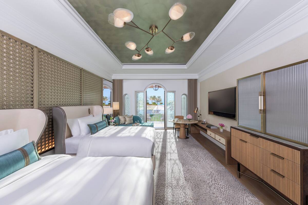 A hotel room with two beds, a TV, a desk, and a view of the outdoors.