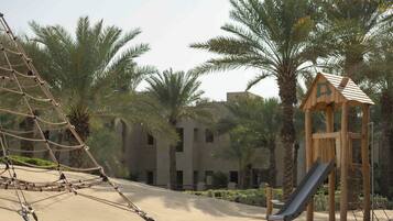 Children can play on the rope climbing structure, slide, and in the wooden playhouse amidst palm trees.