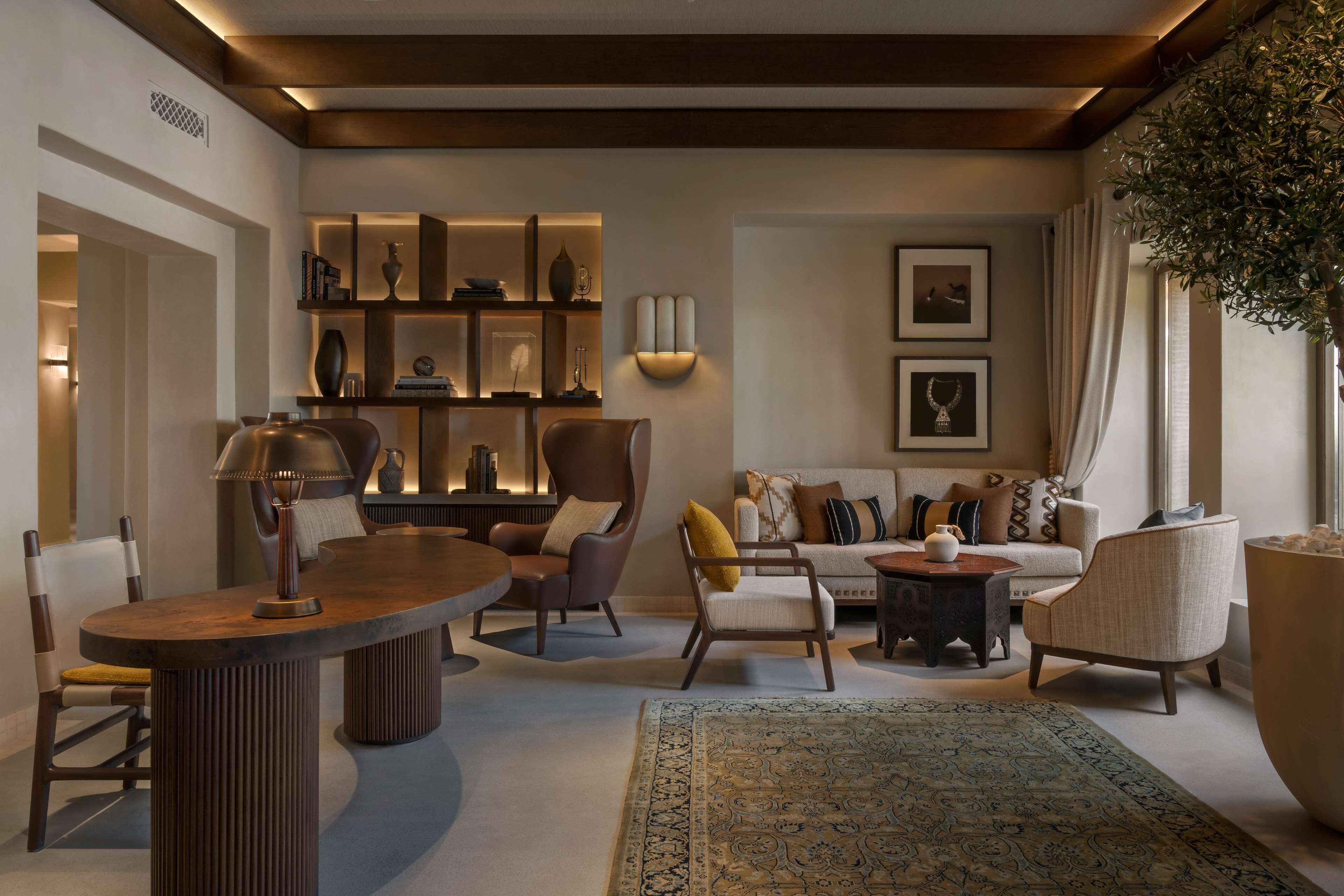 A modern living room with a round wooden table, leather armchairs, a sofa, and a large rug. There is a bookshelf with decorative items and wall-mounted lighting.