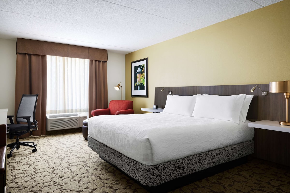 Hotelkamer met een groot bed, een bureau met stoel, een rode fauteuil, een raam met gordijnen en een schilderij aan de muur.
