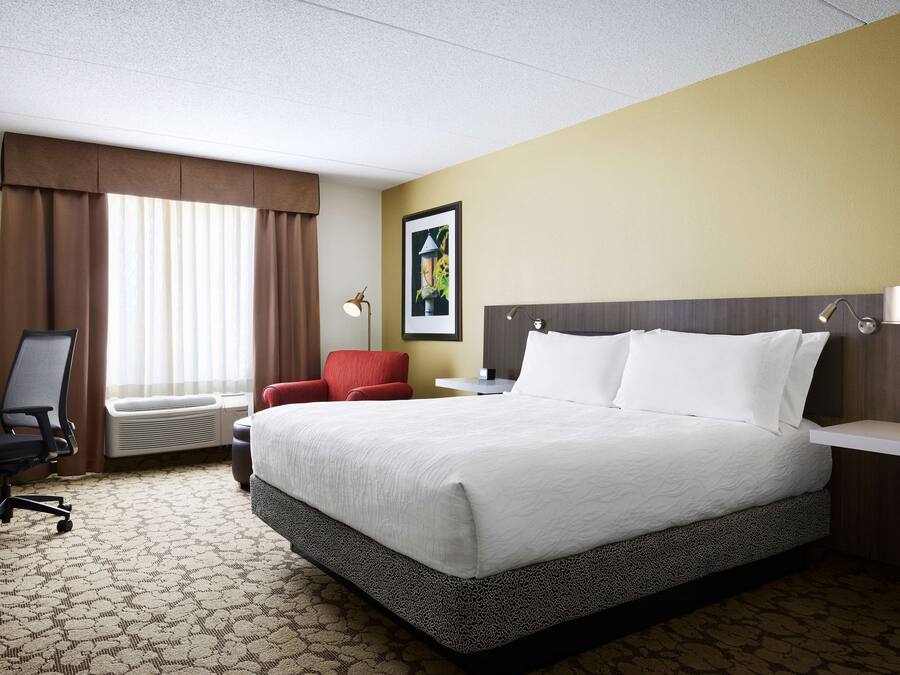 A hotel room with a large bed, a desk with a chair, a red armchair, a window with curtains, and a painting on the wall.