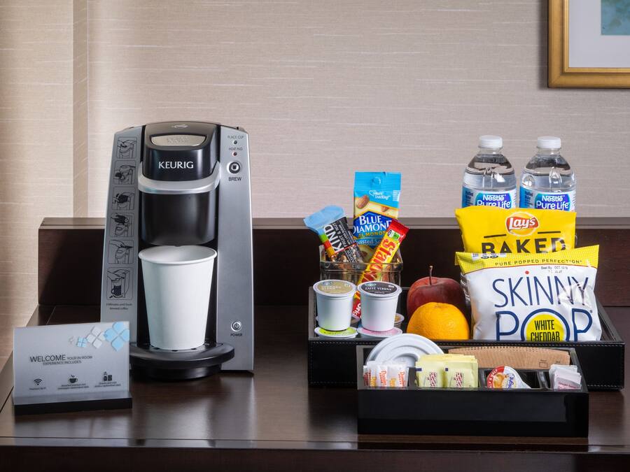 A coffee maker, a welcome sign, snacks, and bottled water on a hotel desk.