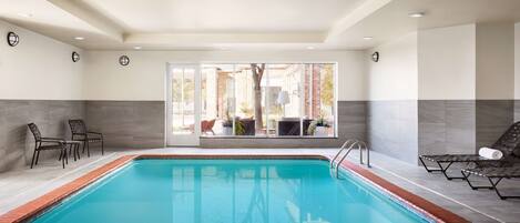 Una piscina con agua azul cristalina, rodeada de sillones de descanso y vista a un edificio de ladrillo a través de amplios ventanales.