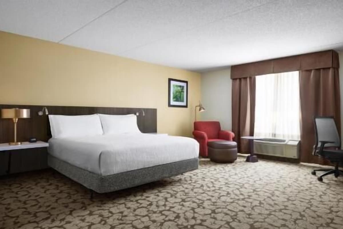 Une chambre d’hôtel avec un lit, un bureau, une chaise, un fauteuil rouge, un tableau au mur et une fenêtre avec des rideaux.