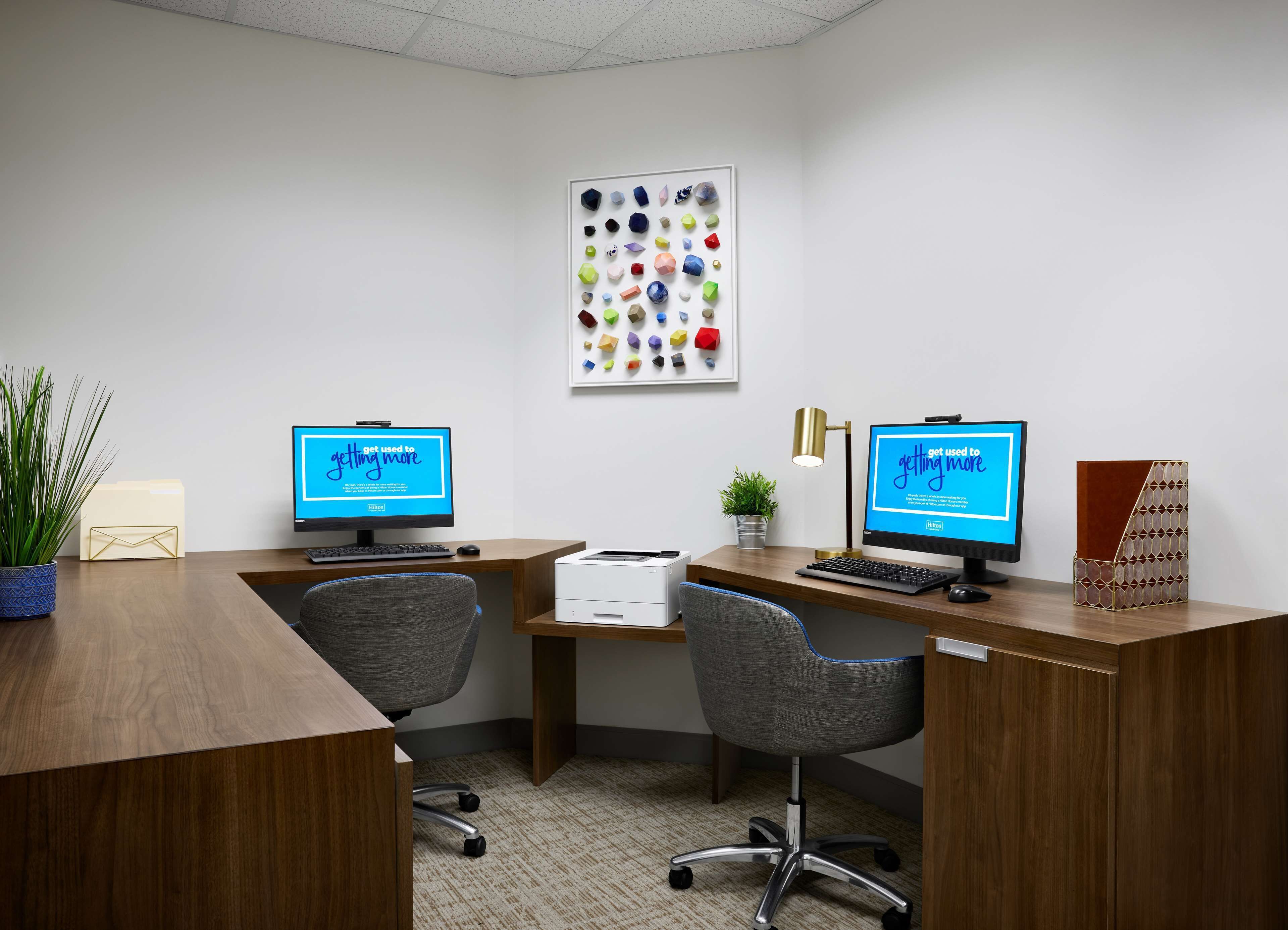 A modern office space with two desks, each with a computer monitor displaying a Hilton advertisement. There is a framed picture of colorful geometric shapes on the wall, a potted plant, and a desk lamp.