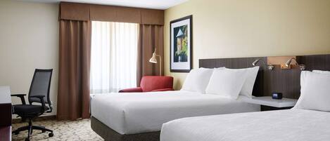 A hotel room with two beds, a desk, a chair, and a lamp.
