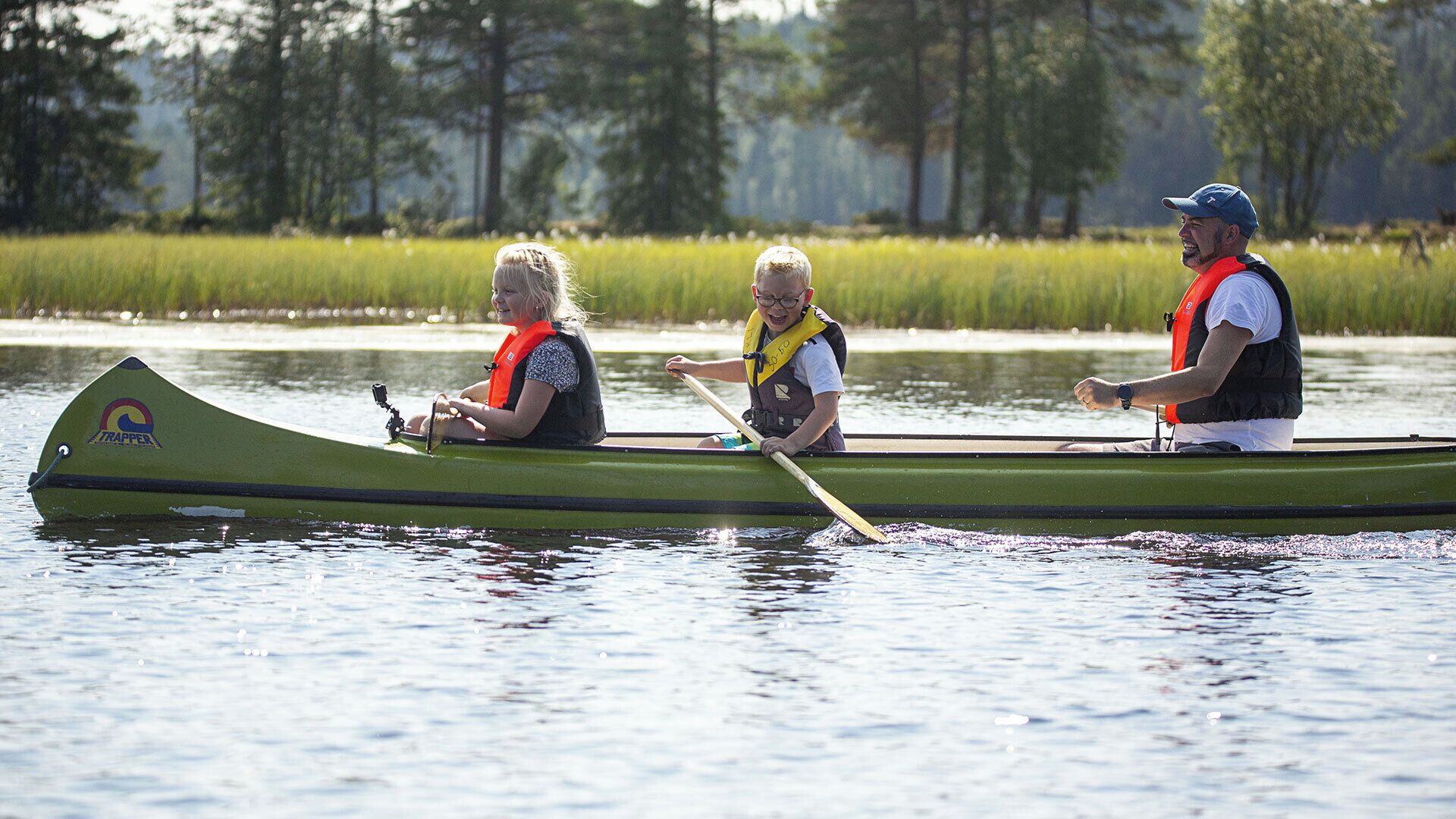 Three people in a green canoe on a calm body of water with trees in the background.