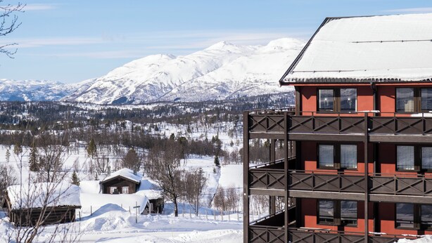 A multi-story building with a red facade and a snow-covered roof, overlooking a snowy mountainous landscape. - Rauland Høgfjellshotell (Vinje)