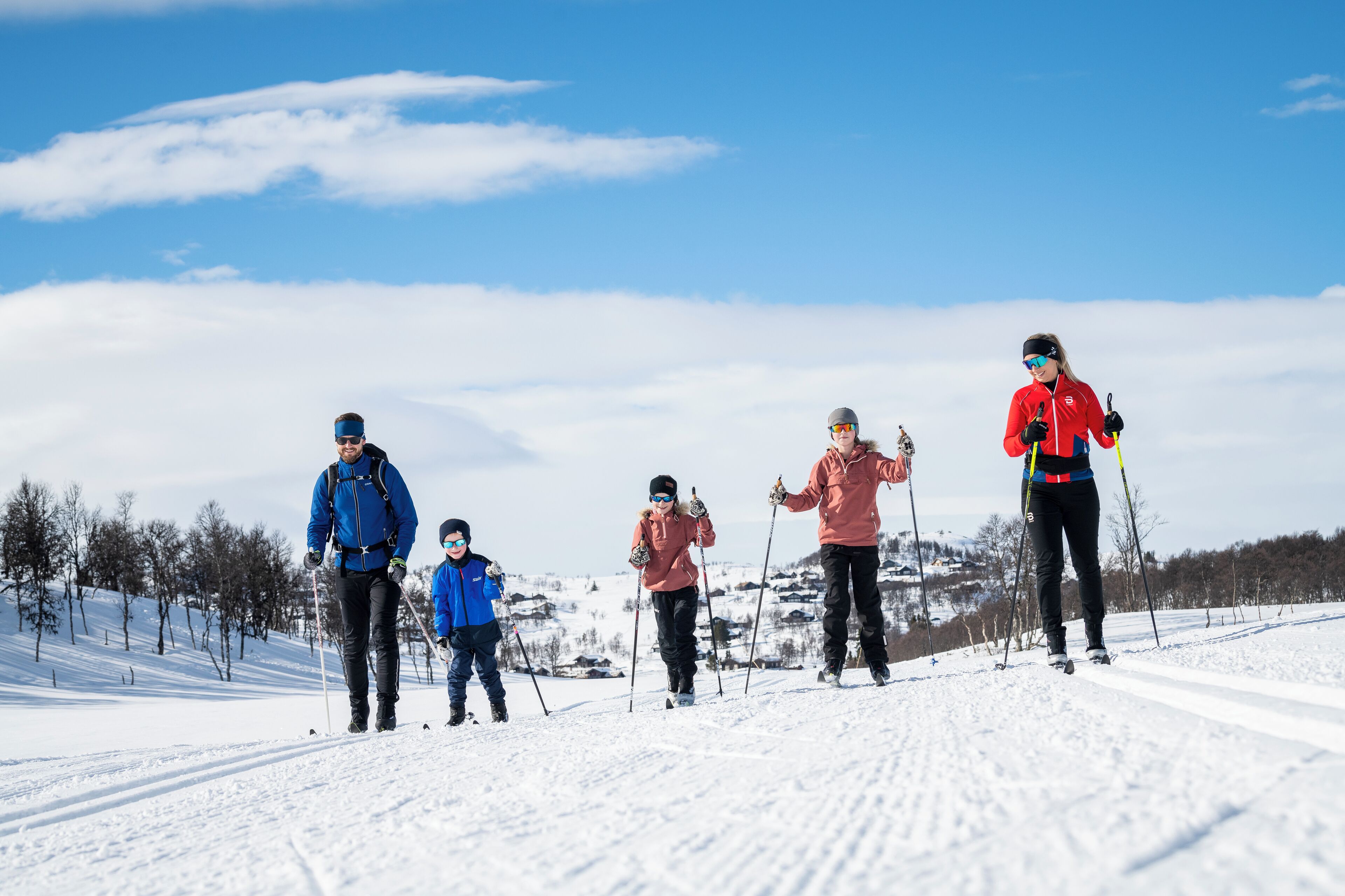 Um grupo de pessoas esquiando de fundo em uma trilha nevada, com árvores e casas ao fundo.