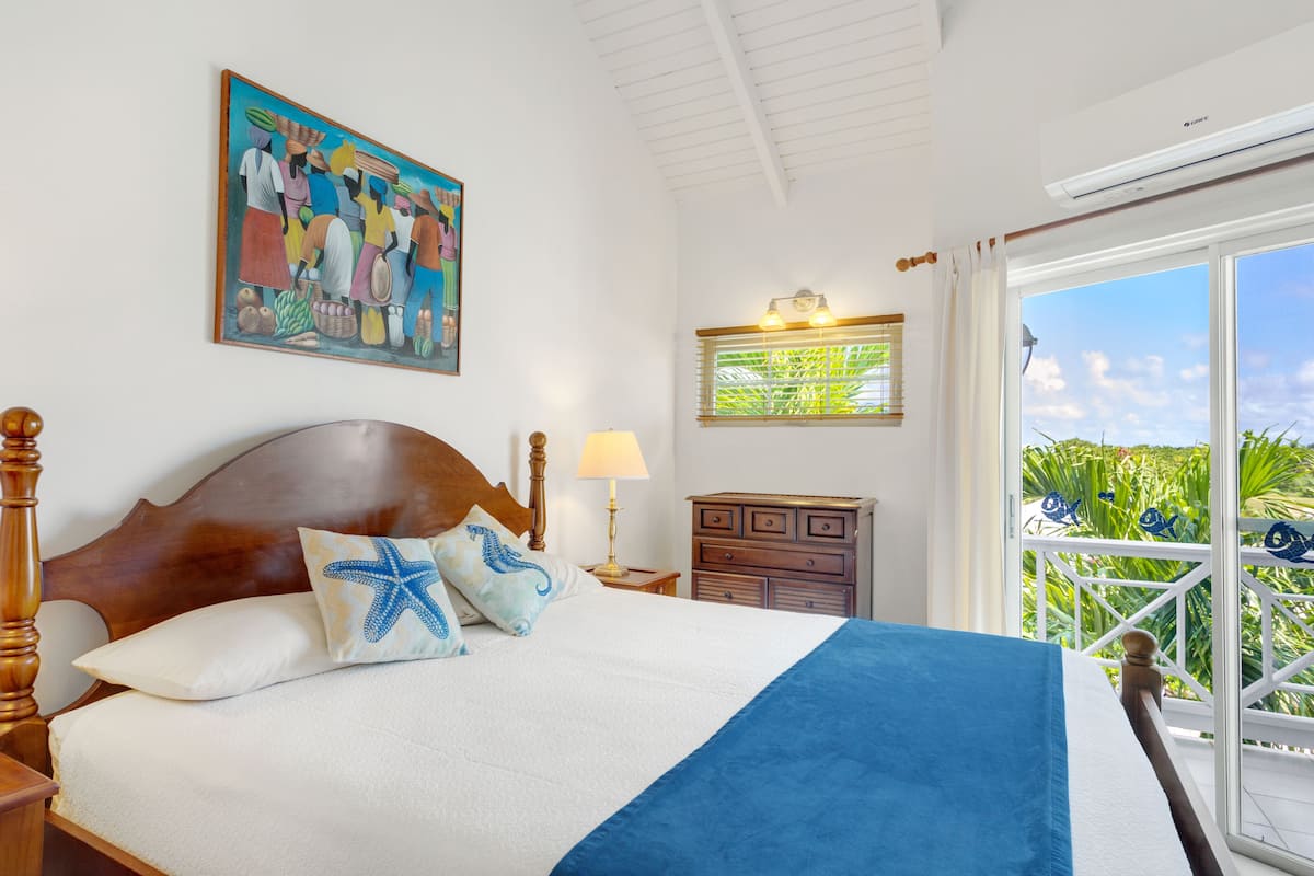 Een slaapkamer met een groot bed, twee bedlampen, een nachtkastje, een dressoir, een schilderij aan de muur en een balkon met uitzicht op groen.