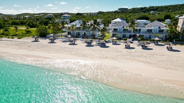 A beachfront property with clear turquoise waters, a sandy beach, and several villas with balconies overlooking the sea.
