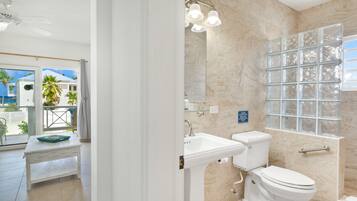 Un baño con inodoro, lavamanos y zona de ducha. Hay un cartel en la pared y una ventana con bloques de vidrio.