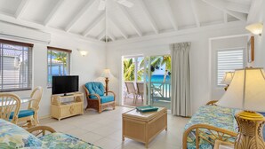 A spacious living room with a large flat-screen TV, wicker furniture, and a view of the ocean through a sliding glass door.