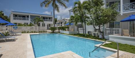 A swimming pool with lounge chairs and umbrellas, surrounded by palm trees and a building with balconies.