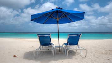 Two white beach chairs under a blue umbrella on a sandy beach with clear turquoise water and a cloudy sky.