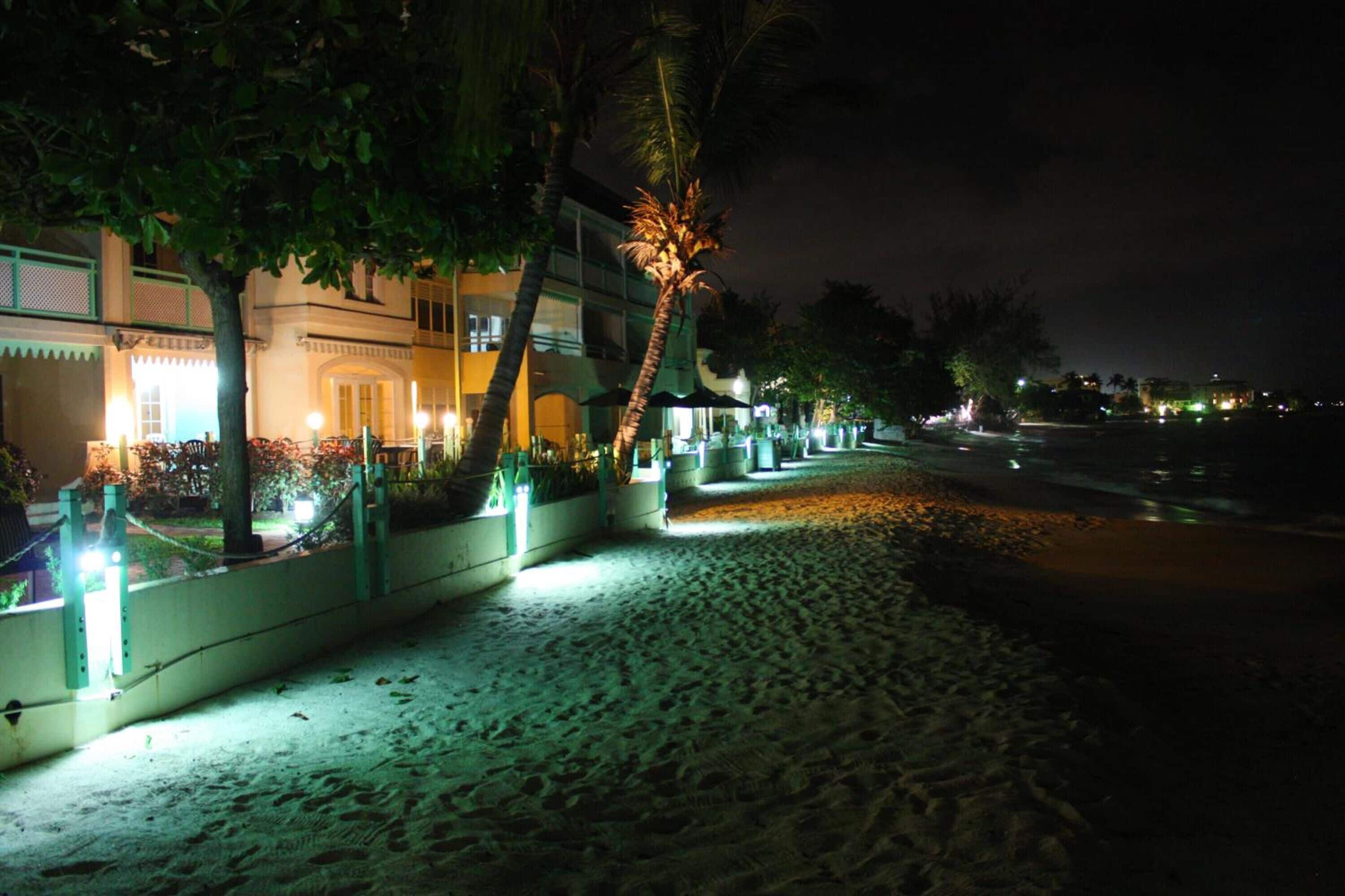 A beachfront hotel with a sandy walkway, palm trees, and a building with illuminated windows.
