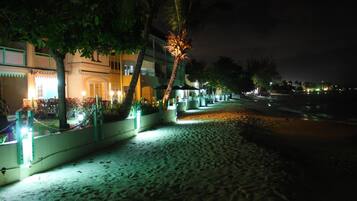 A beachfront hotel with a sandy walkway, palm trees, and a building with illuminated windows.