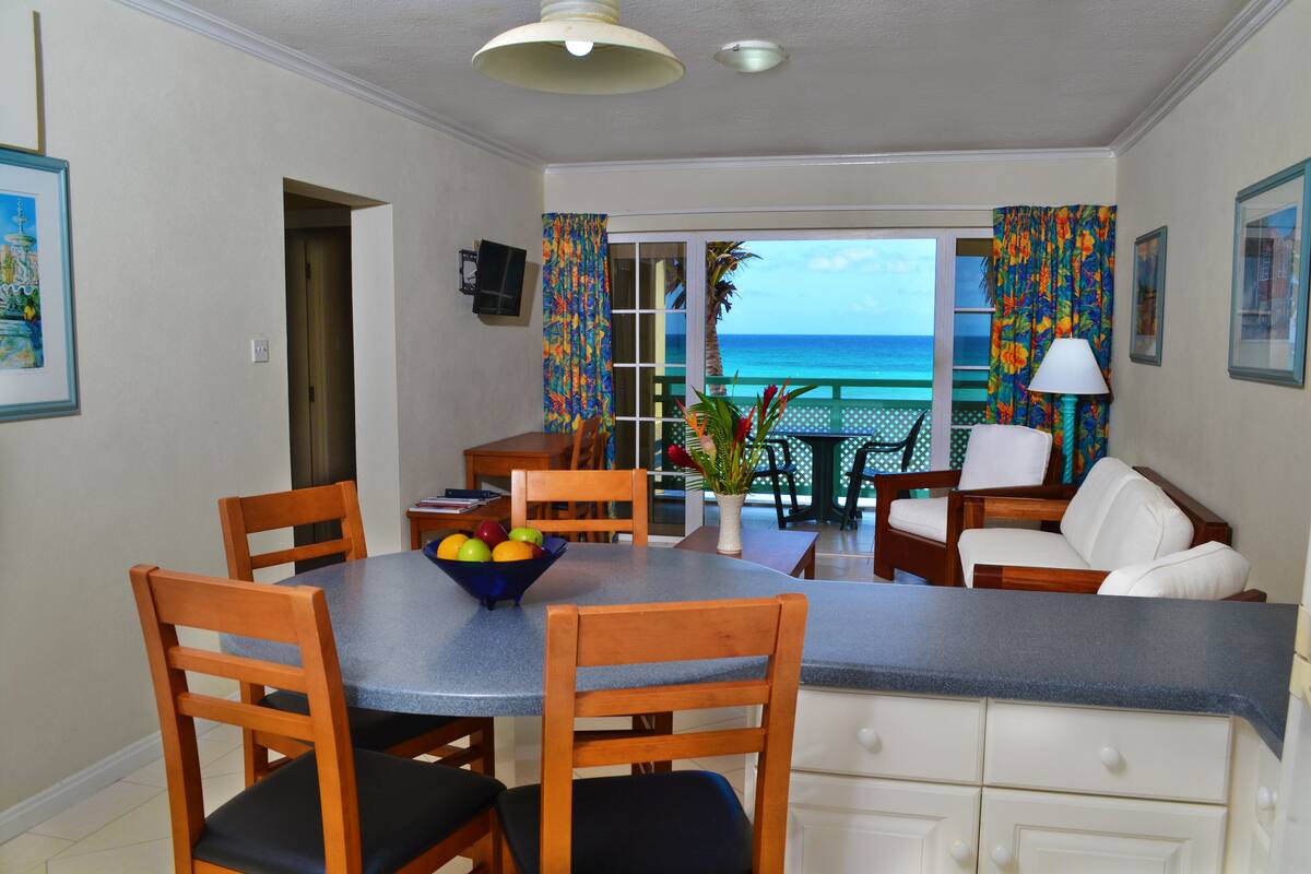 A dining area with a table, chairs, and a bowl of fruit. There is a view of the ocean through the sliding glass door.