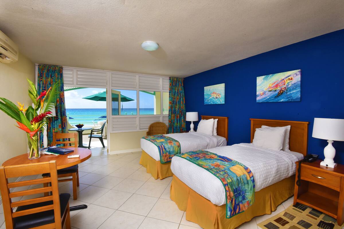 A hotel room with two beds, a dining table, chairs, a small table with a lamp, and a view of the ocean.