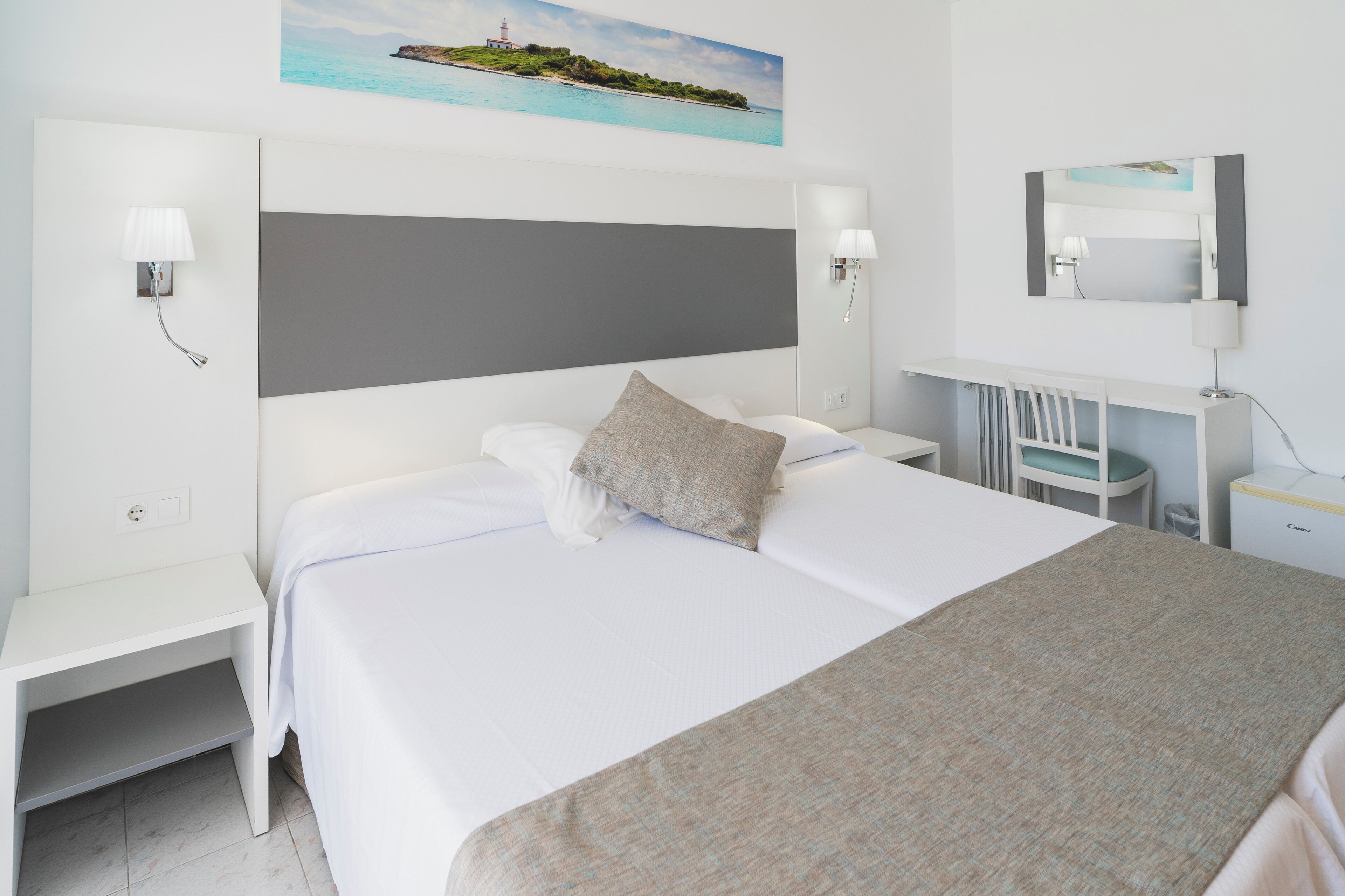 Habitación de hotel con una cama grande, una mesita de noche, un escritorio, una silla, una lámpara de escritorio, un televisor montado en la pared y un cuadro de un paisaje costero.