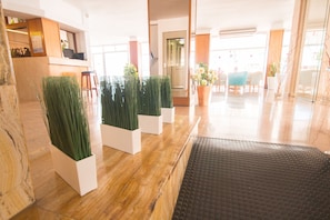 Un espace d’accueil comprenant un comptoir de réception en bois, des jardinières blanches ornées de plantes vertes, un tapis noir à texture, et un coin salon avec des chaises et une table.