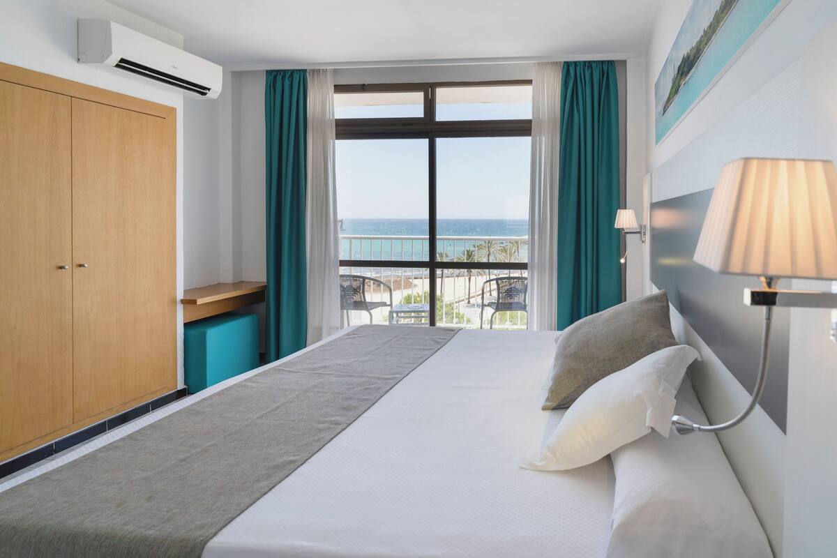Quarto de hotel com uma cama, uma escrivaninha, uma cadeira, uma janela com vista para o mar e um ar condicionado.