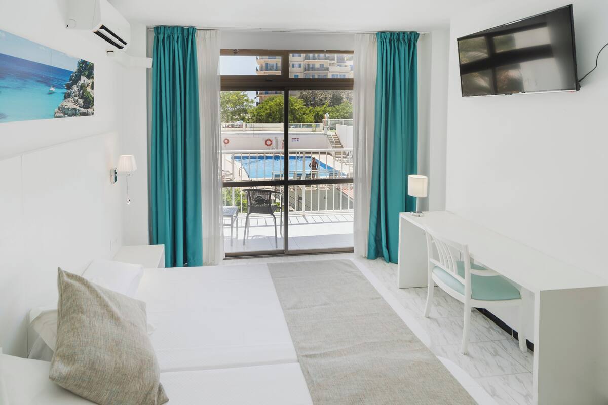 Quarto de hotel com cama, escrivaninha, cadeira, televisão e vista para uma piscina e edifícios.