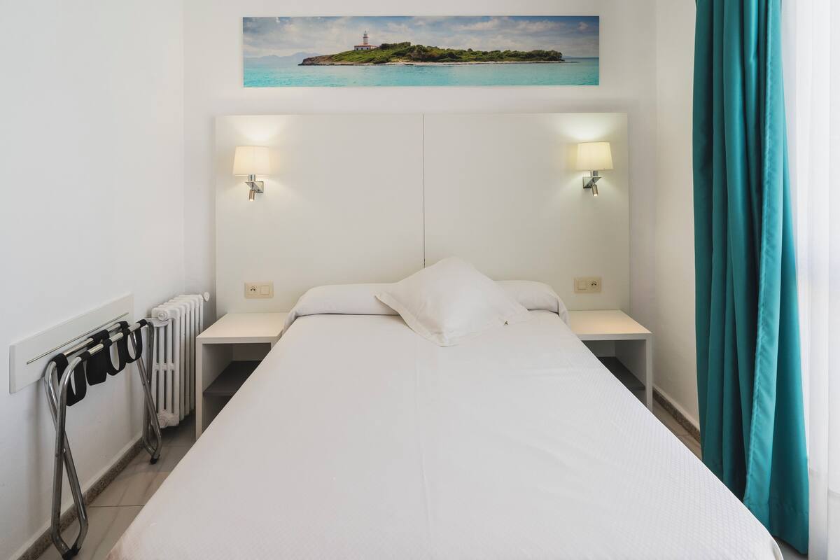 Cama bem arrumada com lençóis brancos, duas mesas de cabeceira com luminárias, um radiador e um porta-toalhas fixado na parede.