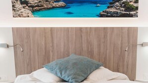 Een slaapkamer met een houten hoofdbord, een bed met wit- en blauw beddengoed, en een schilderij van een kustlandschap erboven.