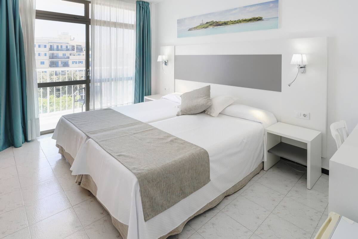 A hotel room with a large bed, a bedside table, a nightstand, a window with curtains, and a painting on the wall.