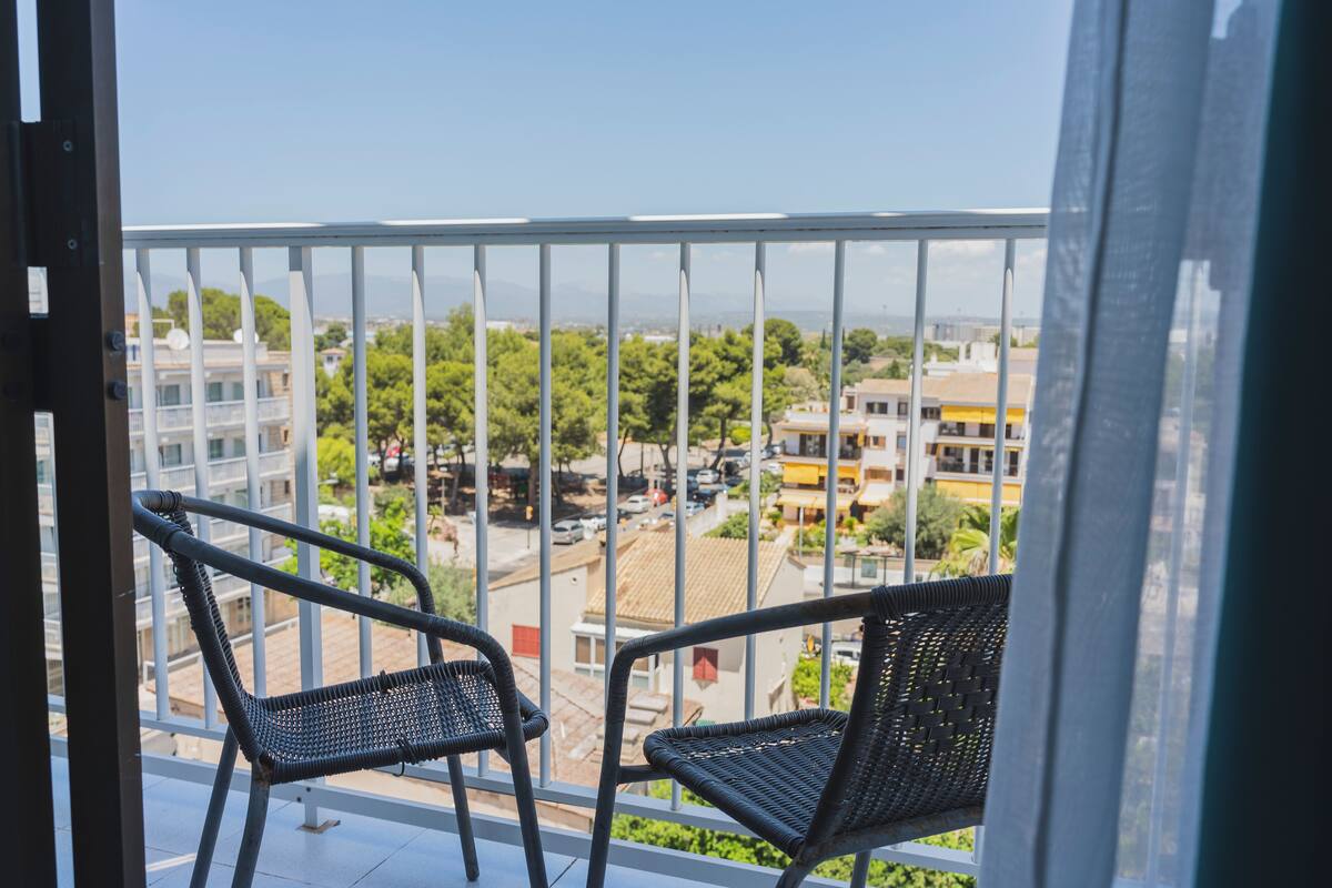 Uma varanda com duas cadeiras de vime, oferecendo vista para um horizonte urbano com edifícios e árvores.