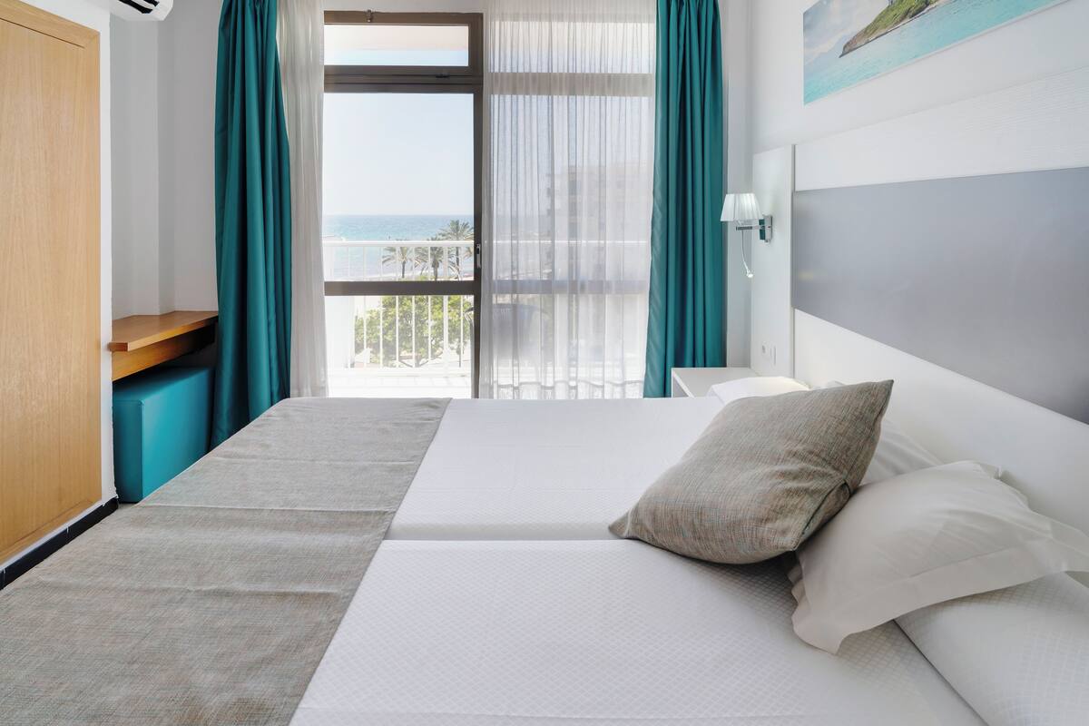 Quarto de hotel com uma cama, mesa de cabeceira de madeira, vista para o mar e janela com cortinas.